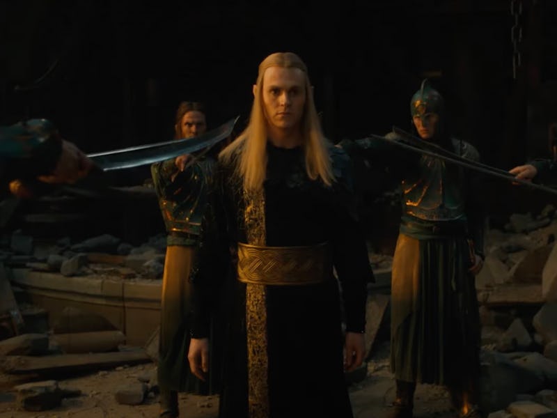 Blonde man in medieval attire, standing with arms outstretched, as swords are pointed at him from mu...